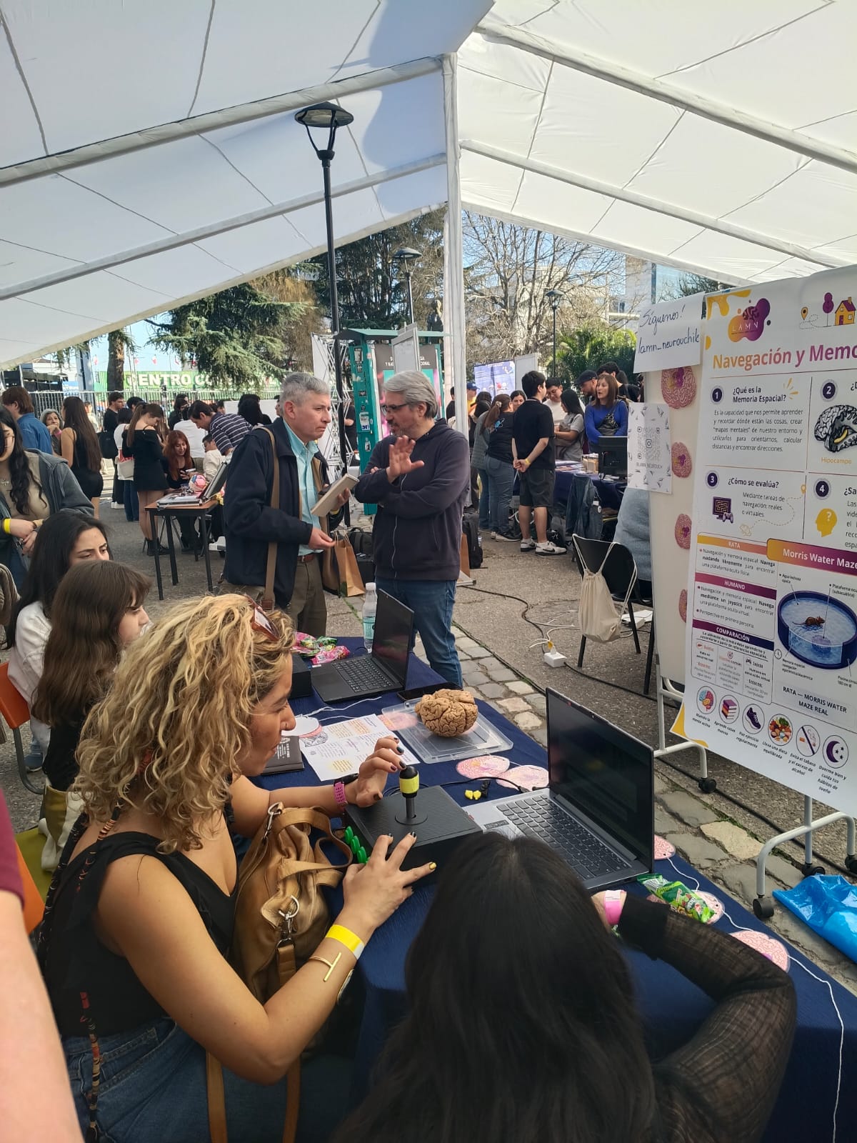 Público jugando con un joystick frente al póster de navegación y memoria espacial en NeuroFest 2026